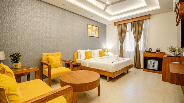 Wide-angle view of a well-furnished hotel bedroom showing a bed, lounge chairs, and large window curtains.