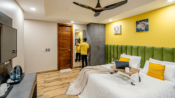 Bright hotel double room featuring a bed with a laptop stand, wooden flooring, a green tufted headboard, and yellow walls.