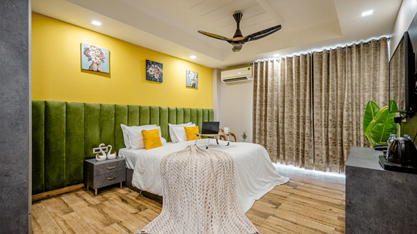 Wide shot of a contemporary hotel room featuring twin beds, green velvet headboards, patterned curtains, and wooden flooring.