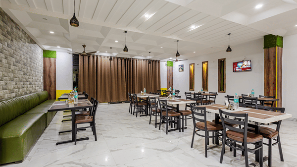 A modern hotel restaurant area with neatly arranged wooden dining tables, dark chairs, and a long green banquette sofa.