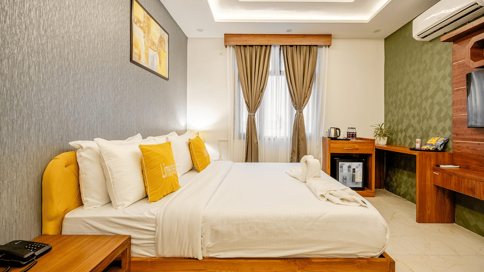 Neatly arranged double bed with yellow pillows next to a wooden bedside cabinet in a hotel room.