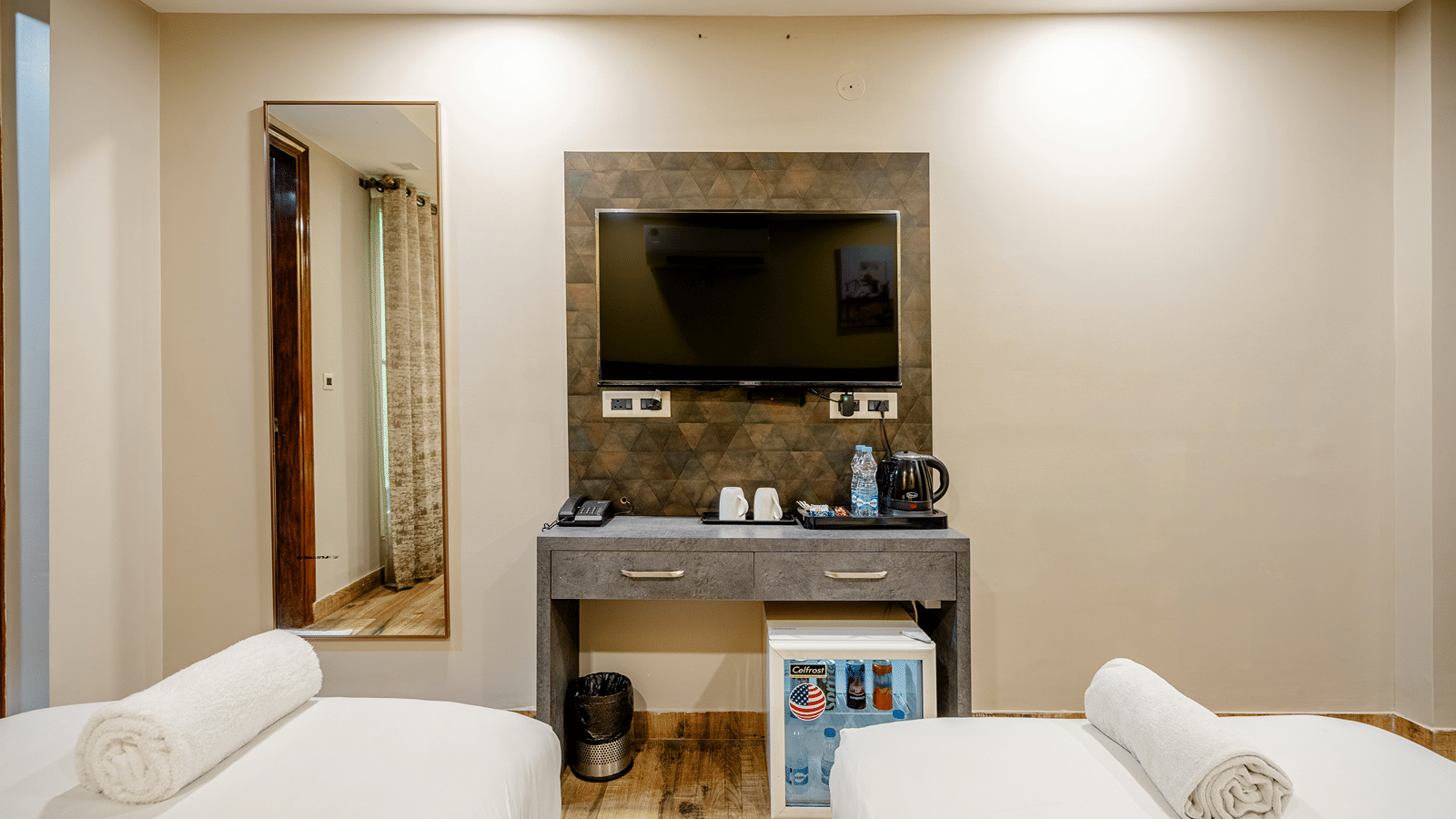 Grey dressing table with a wall-mounted television and a mini-fridge nestled underneath, set between two twin beds.