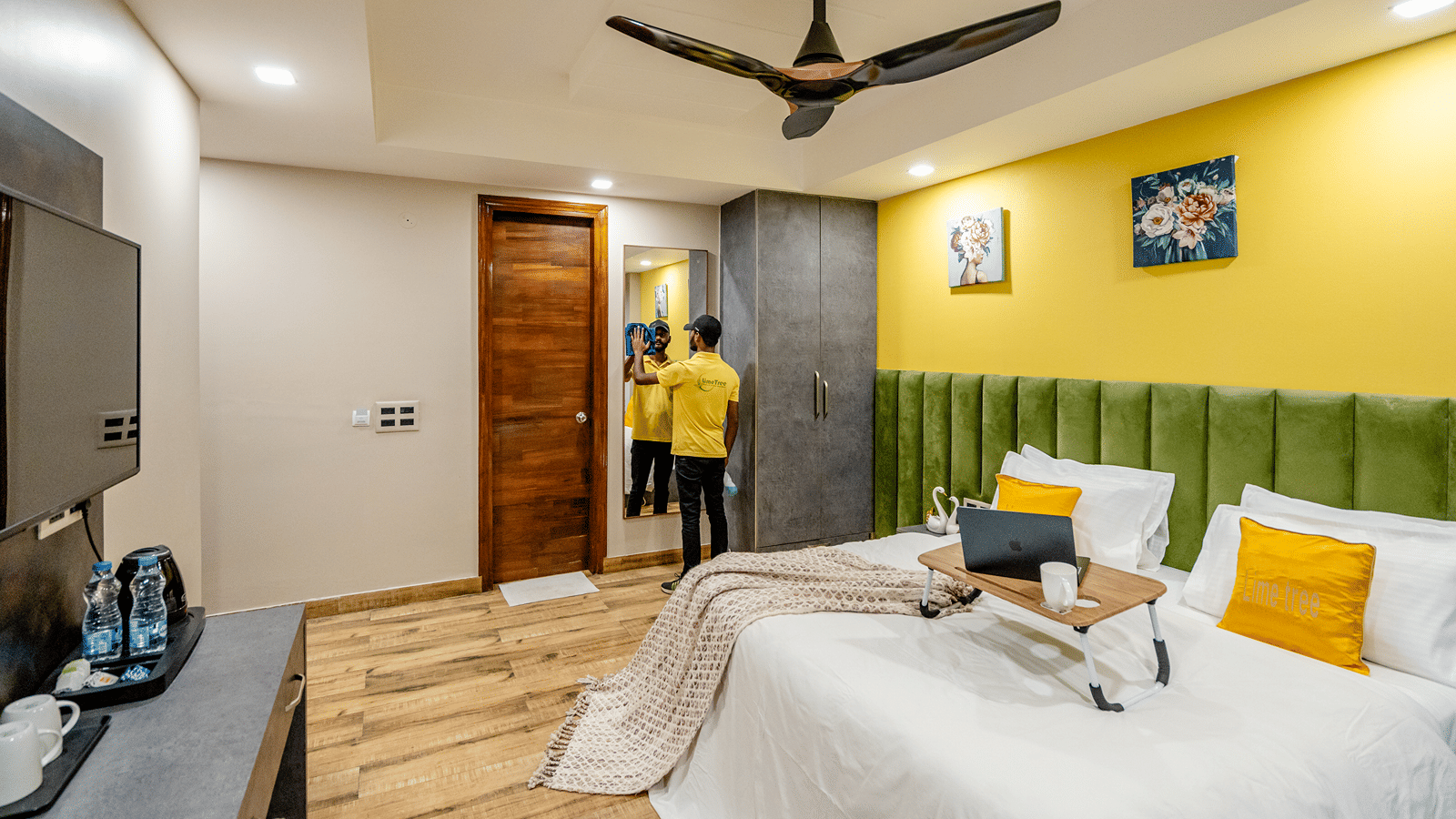 Bright hotel double room featuring a bed with a laptop stand, wooden flooring, a green tufted headboard, and yellow walls.