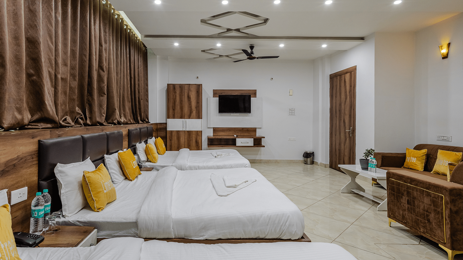A wide view of a spacious hotel family suite with three double beds, a brown fabric sofa, and full-length curtains.