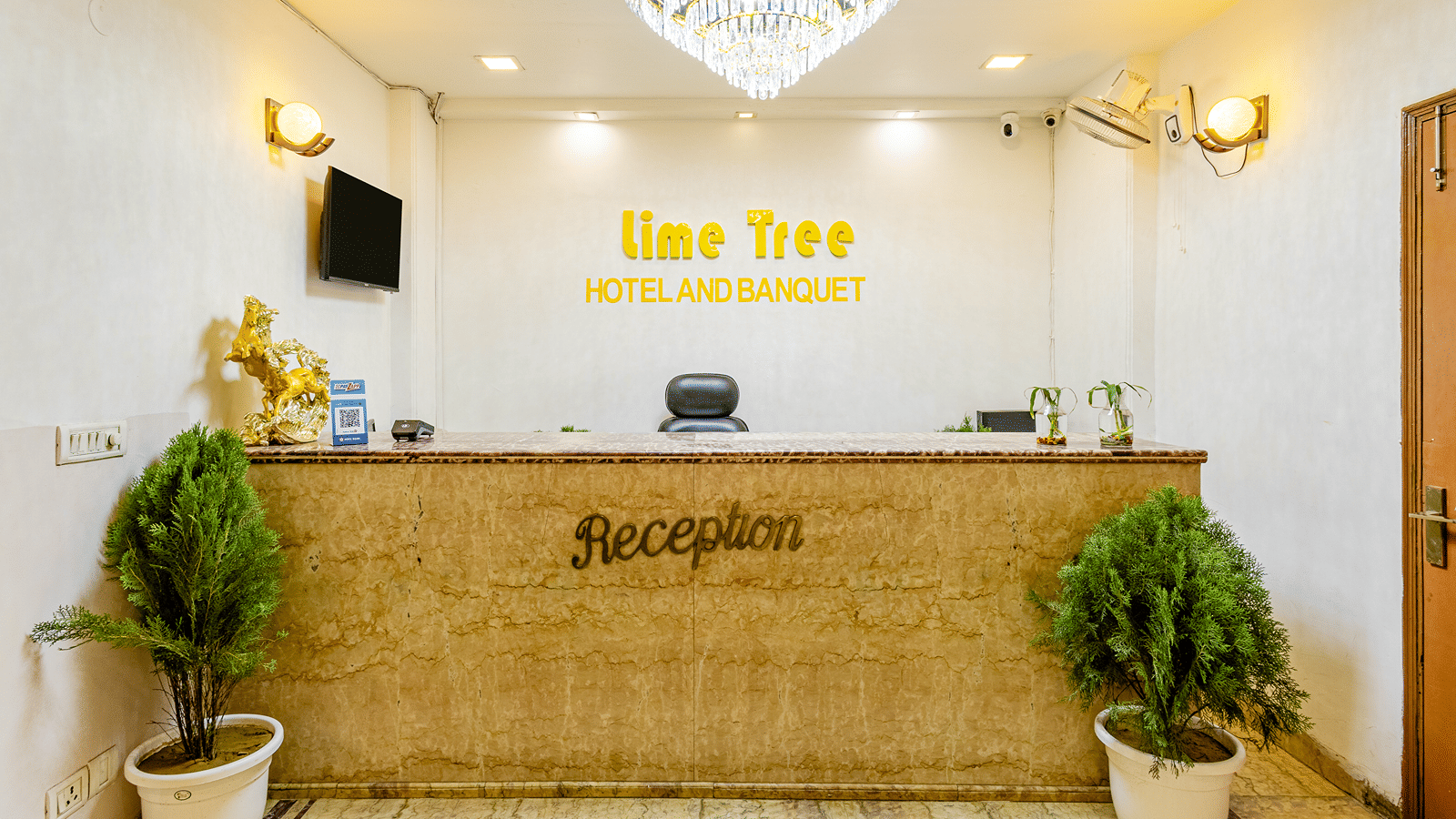 Reception desk of Lime Tree Hotels & Service Apartment Private Limited featuring potted plants and a chandelier lit ceiling.