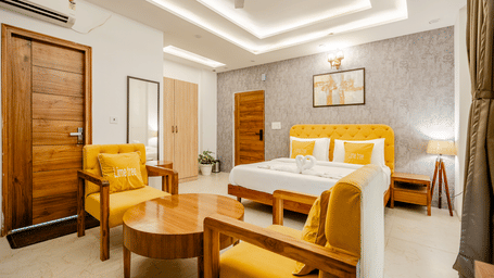 Spacious hotel suite containing a double bed and a wooden seating area with yellow upholstered armchairs.