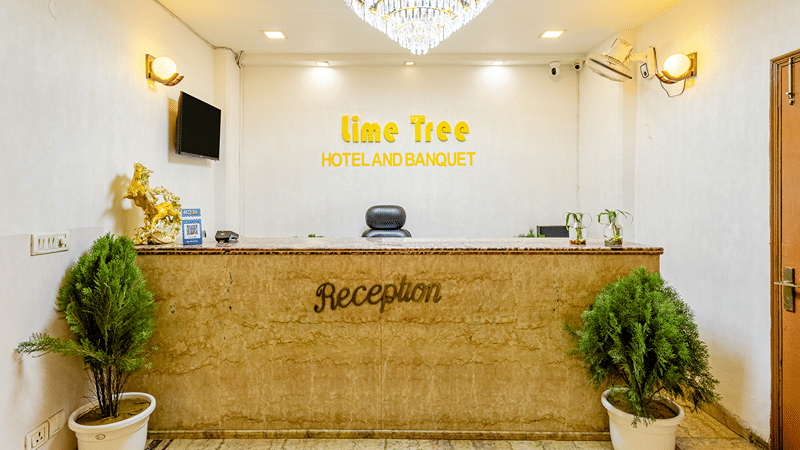 Reception desk of Lime Tree Hotels & Service Apartment Private Limited featuring potted plants and a chandelier lit ceiling.
