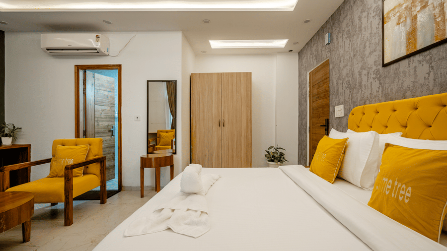 View from the foot of a hotel bed showing a wooden wardrobe, vanity mirror, and yellow accent chairs.