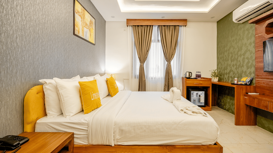 Neatly arranged double bed with yellow pillows next to a wooden bedside cabinet in a hotel room.