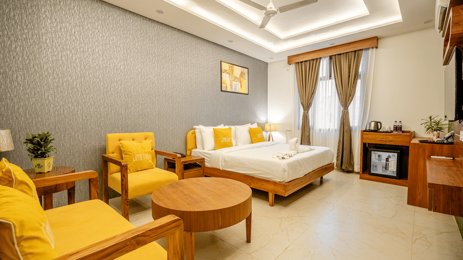 Wide-angle view of a well-furnished hotel bedroom showing a bed, lounge chairs, and large window curtains.