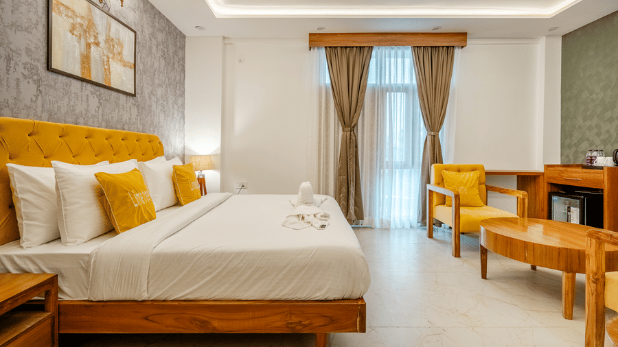 Large hotel room showing a dressed double bed with yellow pillows, floor-to-ceiling curtains, and lounge seating.