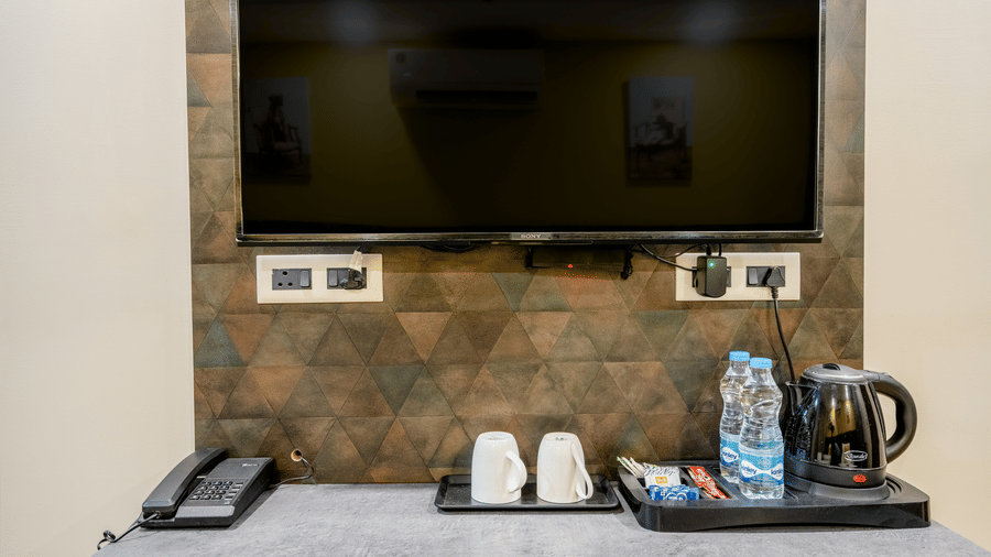 A flat-screen television mounted above a console table equipped with an electric kettle, mugs, and complimentary water bottles.