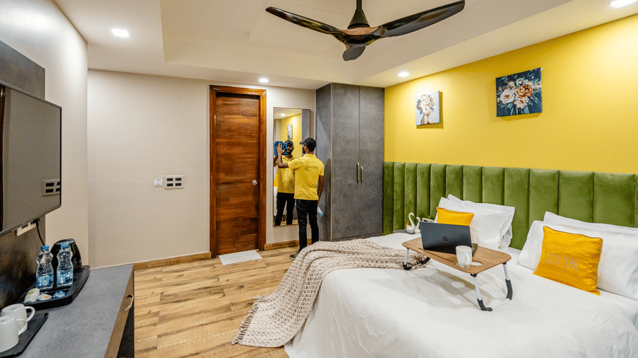 Bright hotel double room featuring a bed with a laptop stand, wooden flooring, a green tufted headboard, and yellow walls.