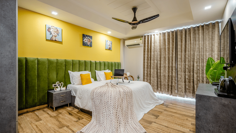 Wide shot of a contemporary hotel room featuring twin beds, green velvet headboards, patterned curtains, and wooden flooring.
