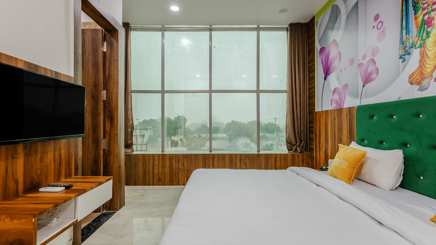 A stylish hotel room showing a white double bed, a green headboard, a wall-mounted TV on a wooden panel, and large windows.