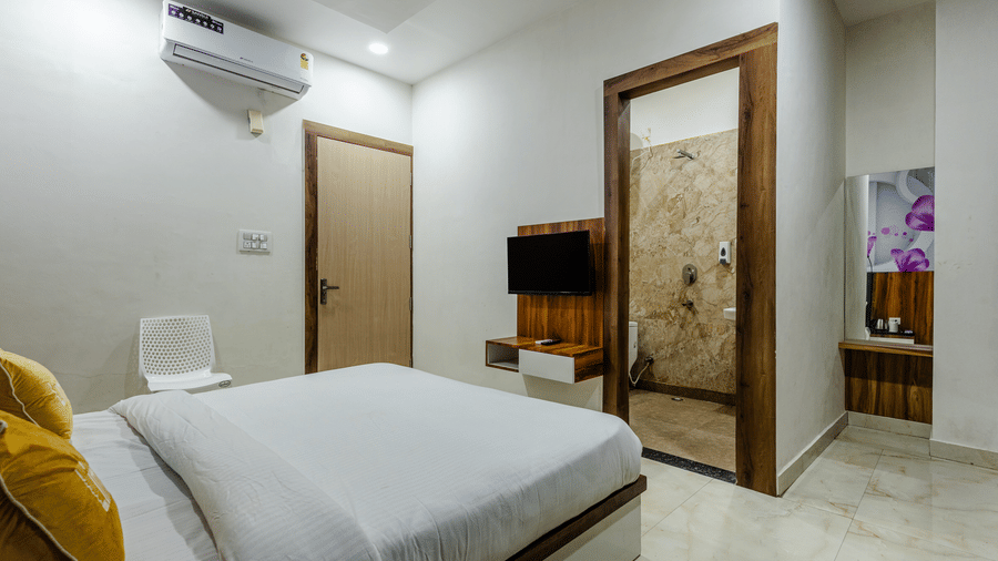A neat double bed in a hotel room with white linen, showing a wall-mounted TV and an open doorway leading to a tiled en suite.