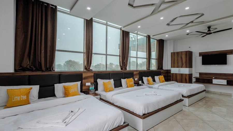 A large hotel room lined with multiple double beds featuring crisp white duvets and yellow cushions by large windows.