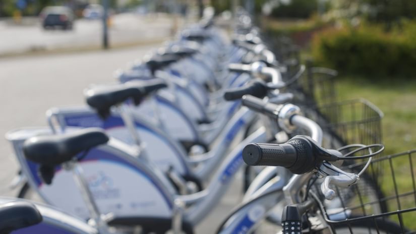 A close up of many cycles parked near a park with a car travelling on the road on the other side in view.