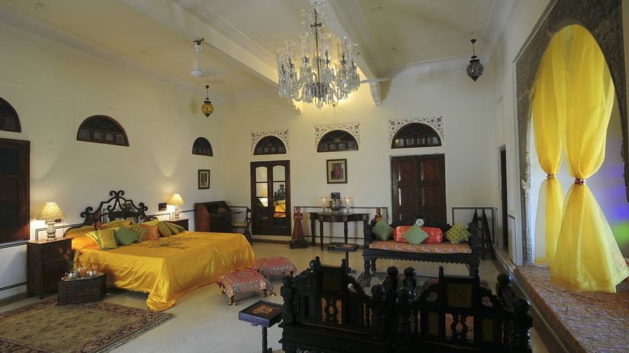 A spacious bedroom at Fort Barli, featuring a vibrant yellow bedspread, a crystal chandelier, and antique furniture.