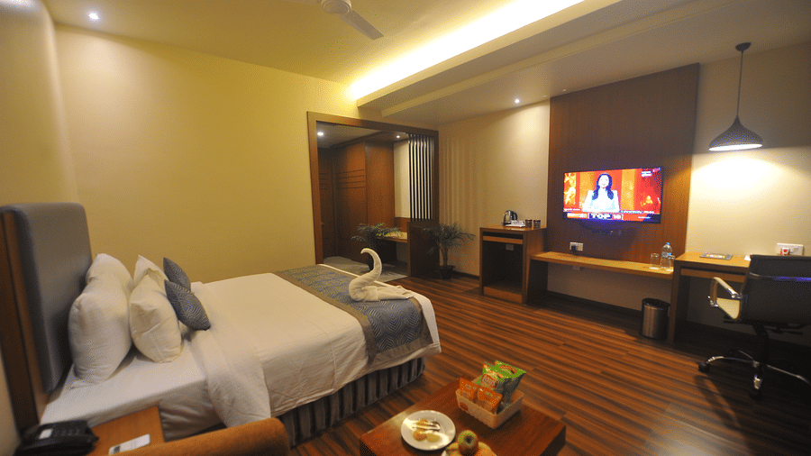 Hotel bed with white pillows and a folded towel arranged decoratively, dark wooden flooring, a wall-mounted television and a fruit tray nearby.