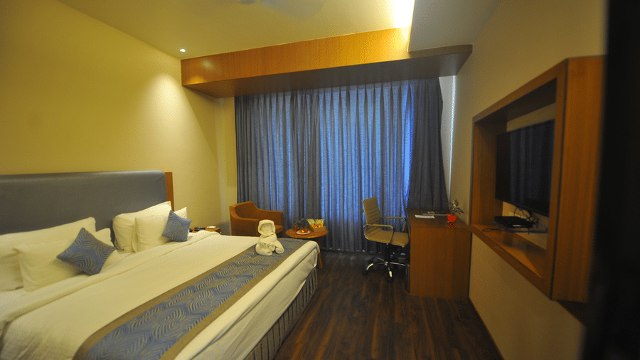 Hotel bedroom with a double bed, long blue curtains, dark wooden flooring, a wall-mounted television and a work desk with a chair placed nearby.
