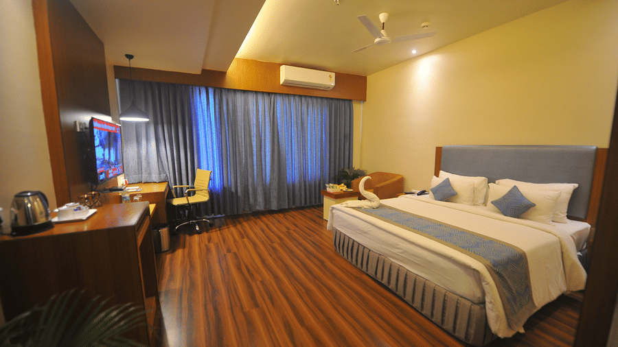 Hotel room with a king-size bed dressed in blue and white linen, dark wooden flooring, a work desk and a wall-mounted television close by it.