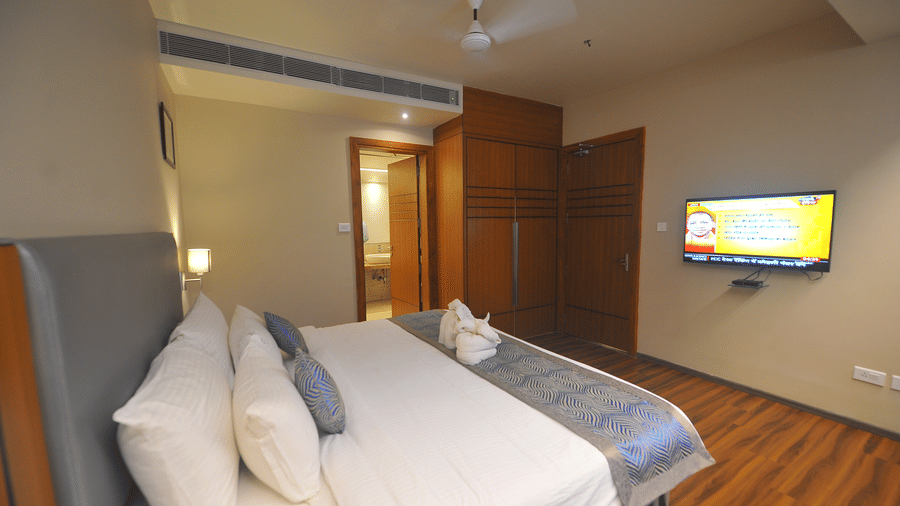 Suite bedroom with a bed dressed in white and blue linen, a grey upholstered headboard, a wall-mounted television and dark wooden flooring.