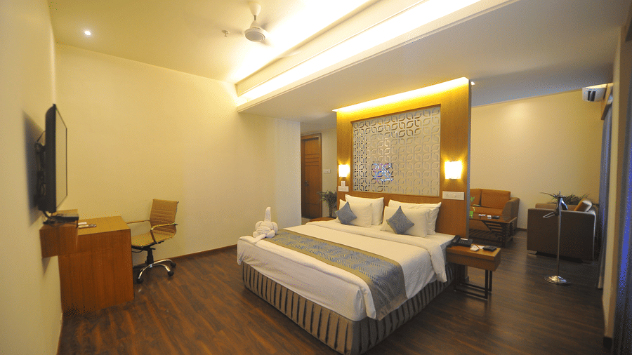 Suite bedroom with a bed, a work desk and chair, a wall-mounted television, warm ceiling lighting and dark wooden flooring throughout the room.