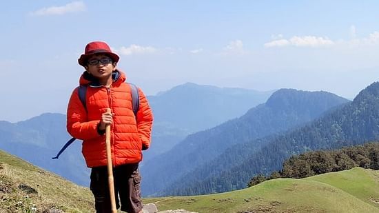 A hiker in a bright red jacket and hat stands on a mountain trail, with a hiking stick and backpack, overlooking a sweeping view of green valleys.