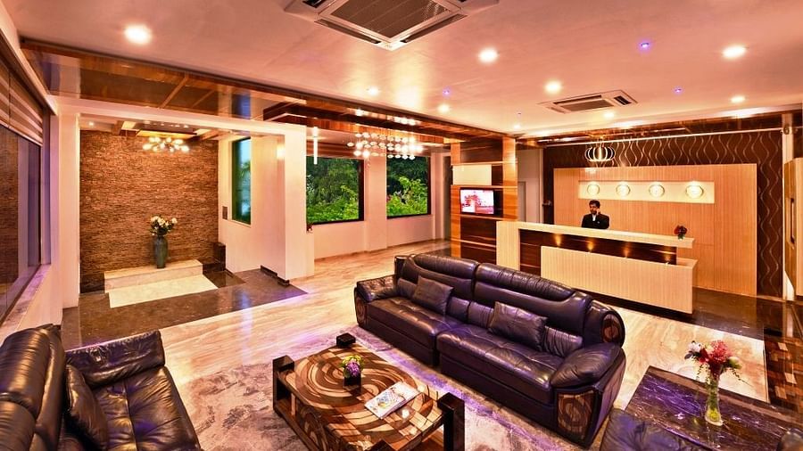 A warmly illuminated hotel lobby, possibly Lords Inn Porbandar, shows comfortable sofas, decorative tables, and a reception desk area.