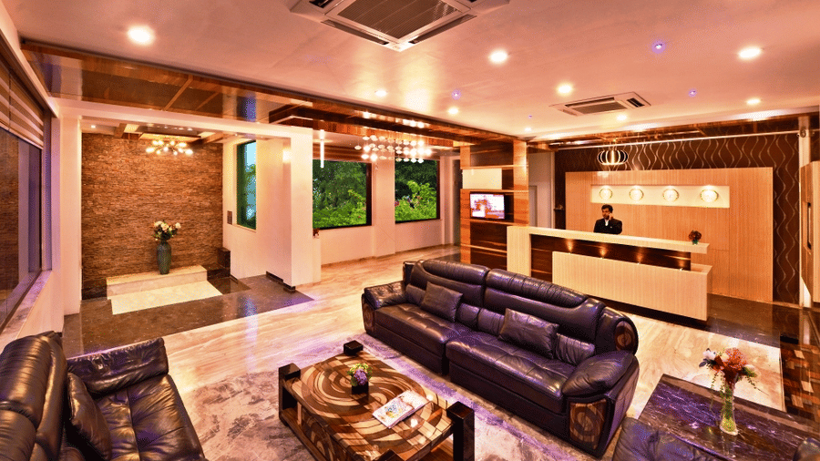 A warmly illuminated hotel lobby, possibly Lords Inn Porbandar, shows comfortable sofas, decorative tables, and a reception desk area.