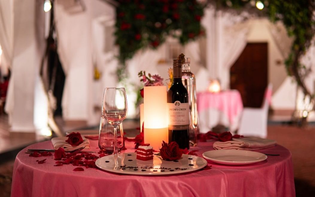 A romantic dining arrangement with a candle and a bottle of wine placed on a table with a dark pink table cloth - Saraca Hotel, Lucknow,