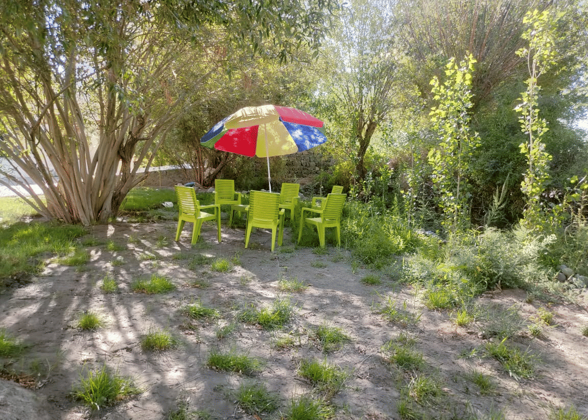 A shaded outdoor seating area with green chairs and a multi-coloured parasol set amongst trees at Indus Serenity Retreat.
