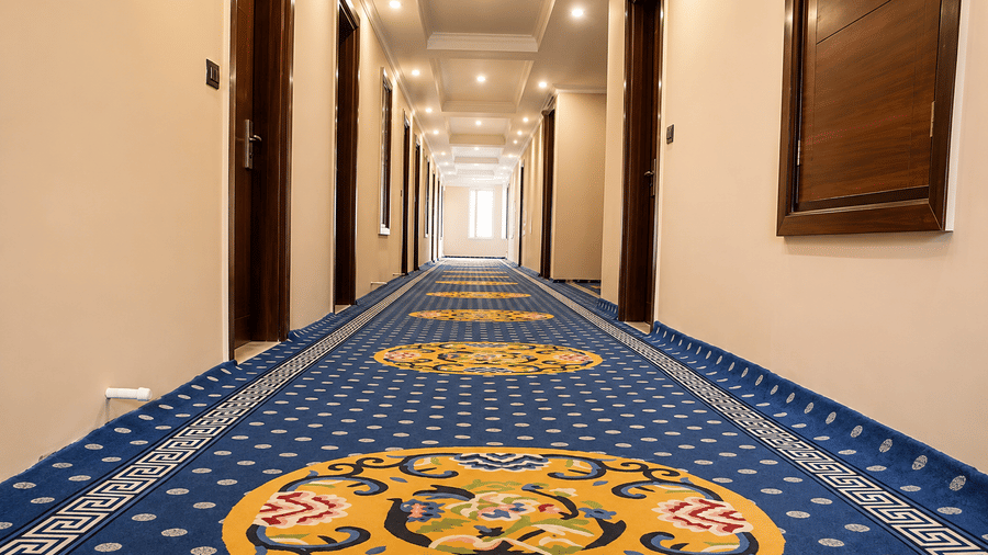 Hotel corridor with a patterned blue carpet featuring traditional motifs, wooden doors on either side and warm ceiling lighting throughout.
