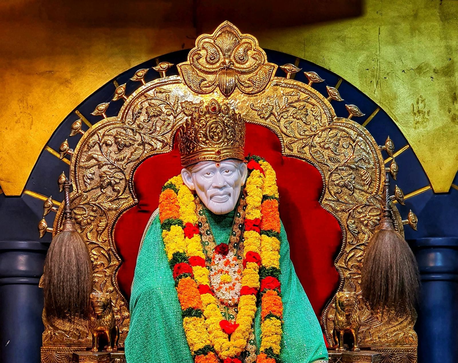 A gold statue of Shri Sai Baba with a yellow and green robe and a yellow flower garland on a throne.