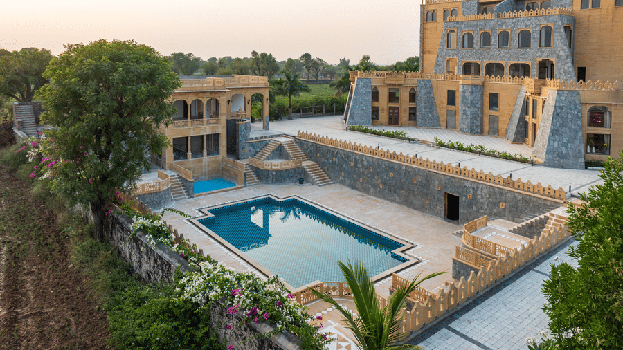 A wide view of the pool area at EsthereaRaj Leela, Ranakpur, featuring a multi-story stone building nestled among trees and landscape.