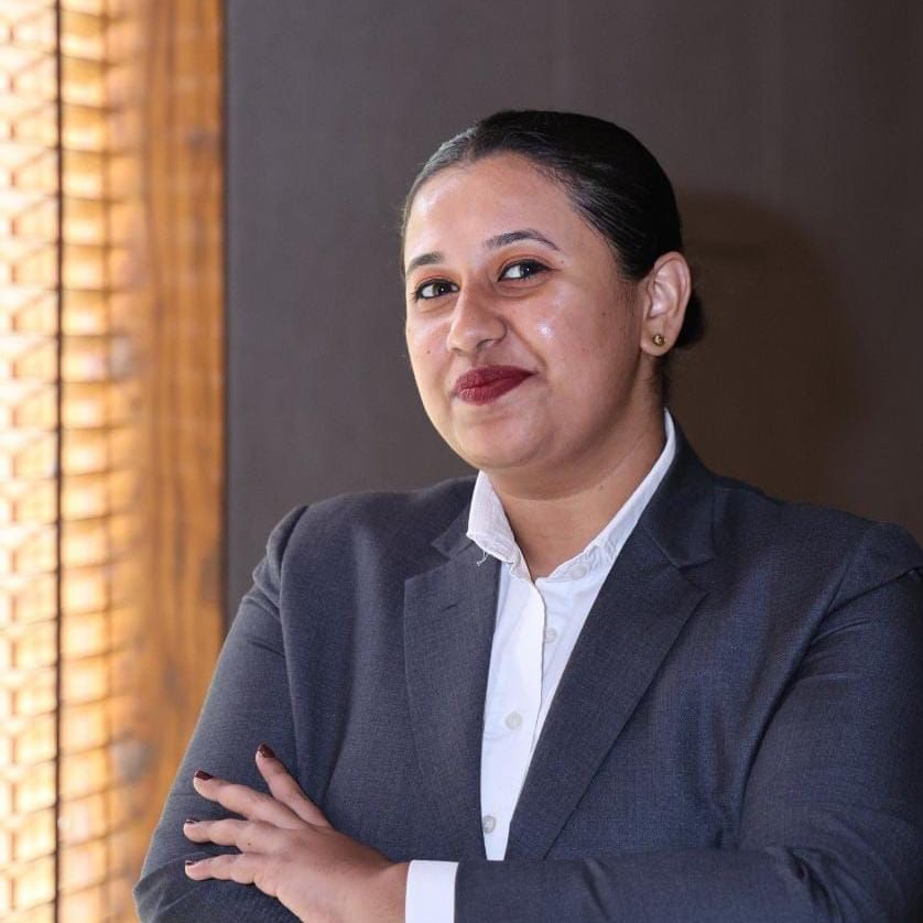 A shot of a smiling woman wearing a grey blazer above a white formal shirt, posing with her hands in a fold | Vedic Village Spa Resort