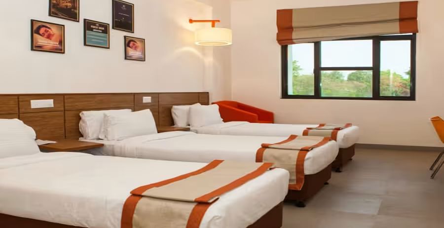 A bright, well-furnished hotel room with three neatly made beds, wall art, and a window- Max Hotel Prayagraj.