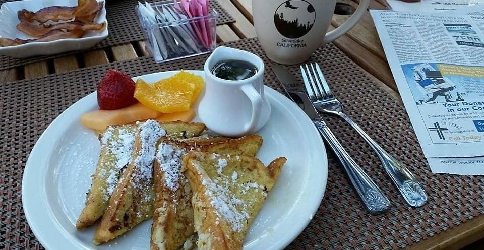 A plate of two churros dusted with powdered sugar, served with fruit and a dipping sauce, alongside cutlery on a patterned placemat
