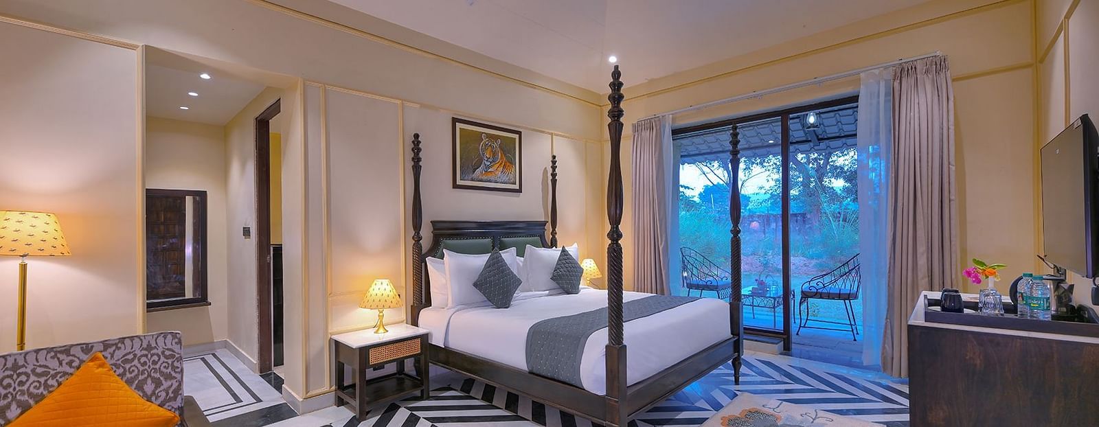 Luxury Suite Cottage with a four-poster bed, a living area with a patterned sofa and orange pillow, black and white chevron flooring, and a balcony - Singhvilas, Ranthambore