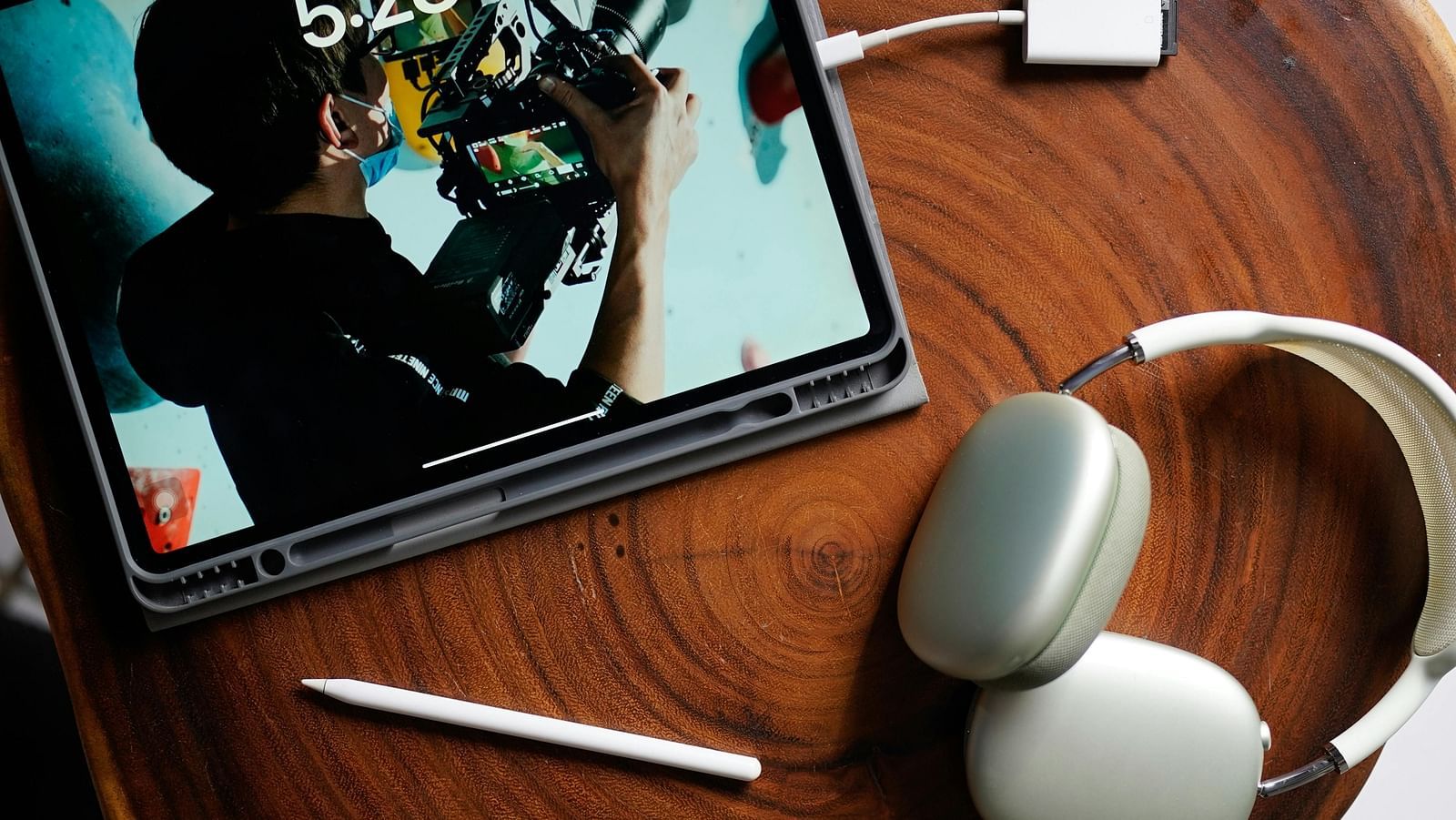 A tablet placed on a wooden surface with headphones, a stylus, and a charging adapter.