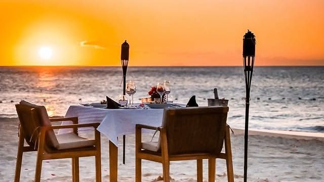 A romantic beach dining setup at sunset with a table and chairs on the sand, illuminated by torches.