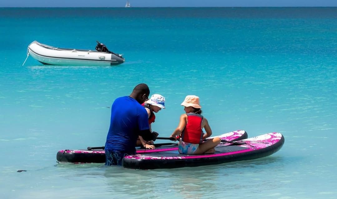 A man and two kids on paddleboards in shallow turquoise water, with an inflatable boat and sailboat in the background.