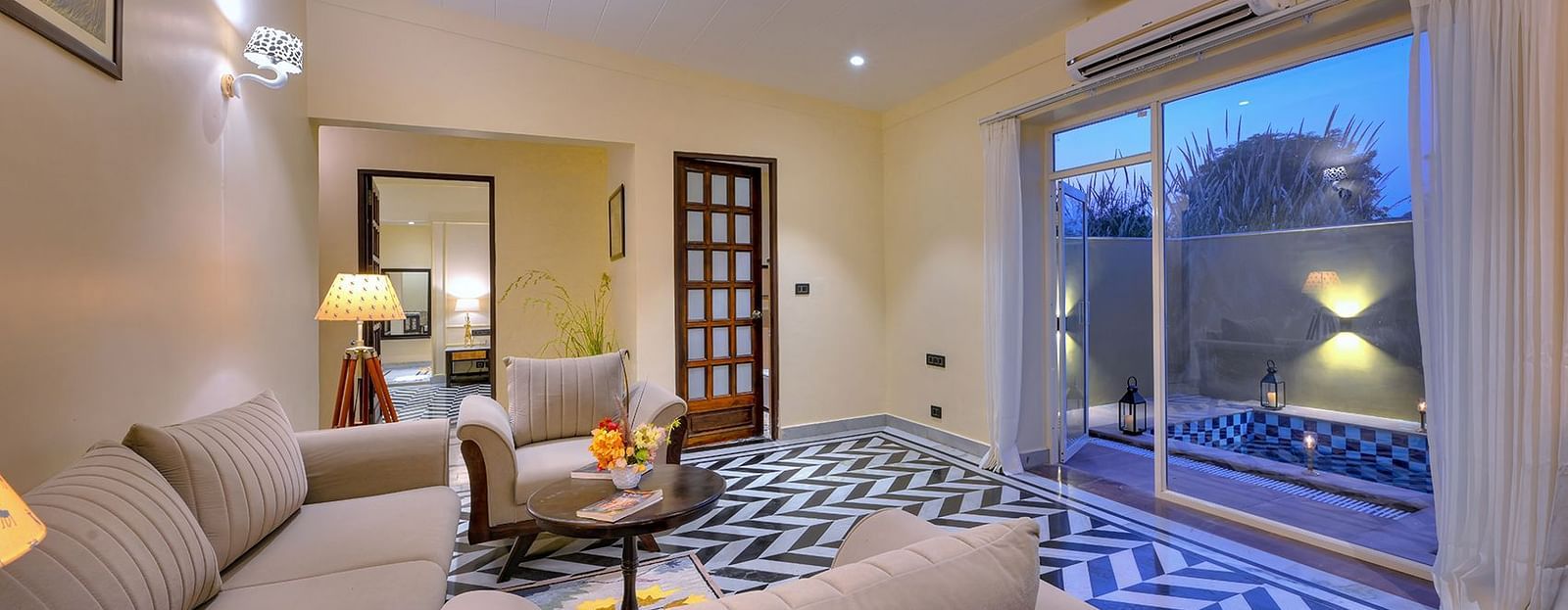 Cosy living room of Premium Suite Cottage with beige coloured sofas, a round coffee table, and patterned flooring, featuring large windows and serene evening lighting - Singhvilas, Ranthambore
