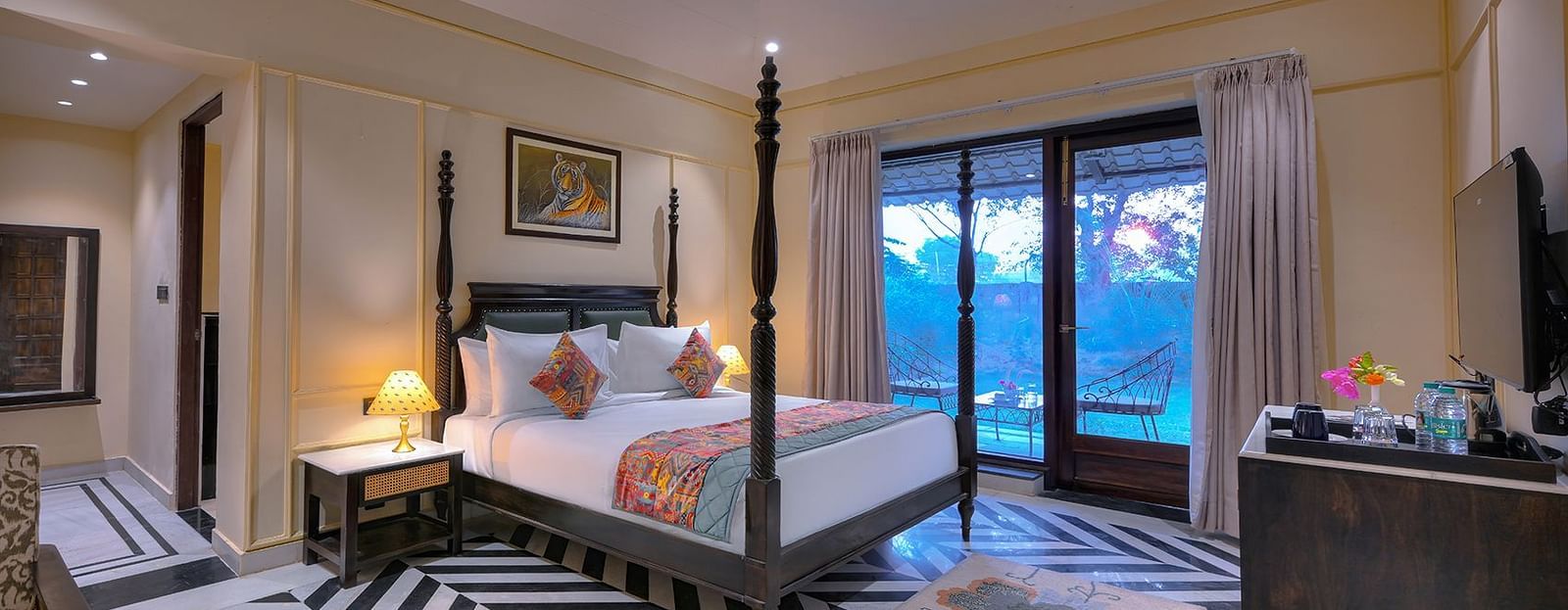 Luxury Suite Cottage featuring a four-poster bed with white linens, a colourful patterned runner and pillows, a patterned armchair, a wall-mounted TV, and sliding glass doors leading to a balcony - Singhvilas, Ranthambore