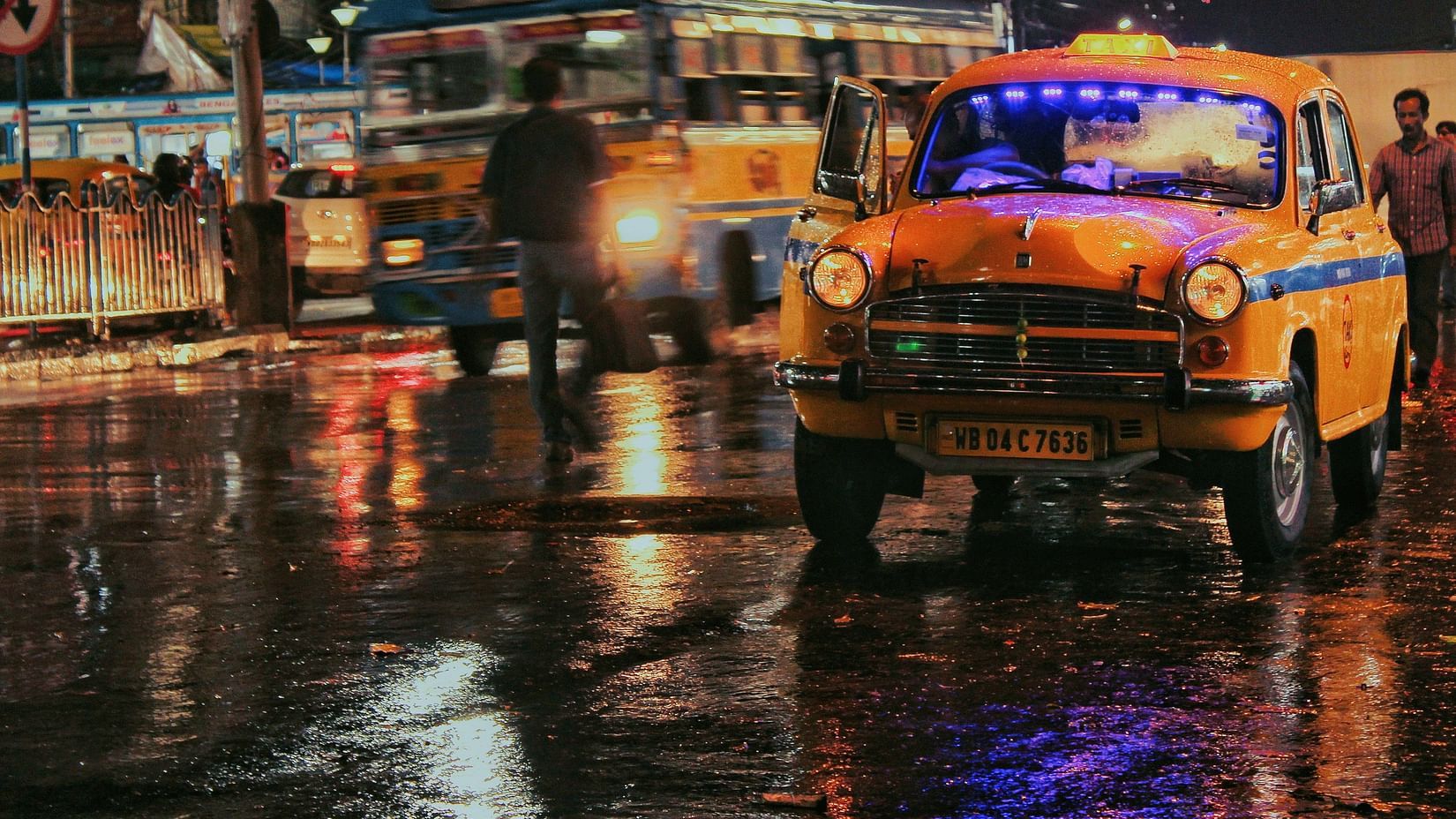A vintage yellow taxi driving through a rainy city street at night with reflections on the wet road.