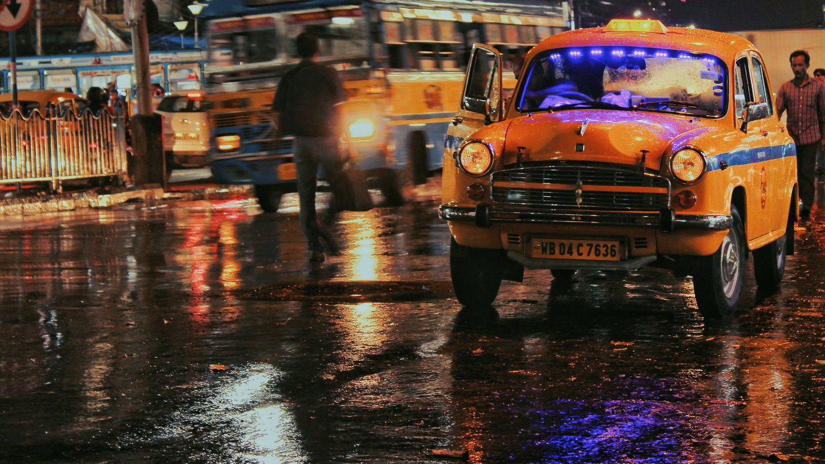 A vintage yellow taxi driving through a rainy city street at night with reflections on the wet road.