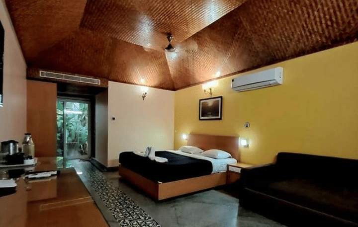 A cosy room with a queen bed, a sofa, wooden furnished ceiling and table with marble floor at Paradise Lagoon Resort, Udupi.