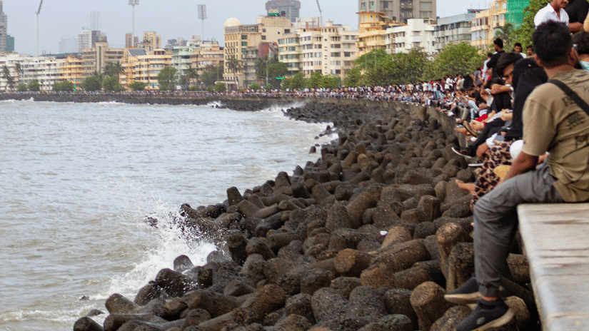 Visitors sit along the rocky shoreline at Marine Drive, enjoying calm monsoon views across Mumbai's waterfront.