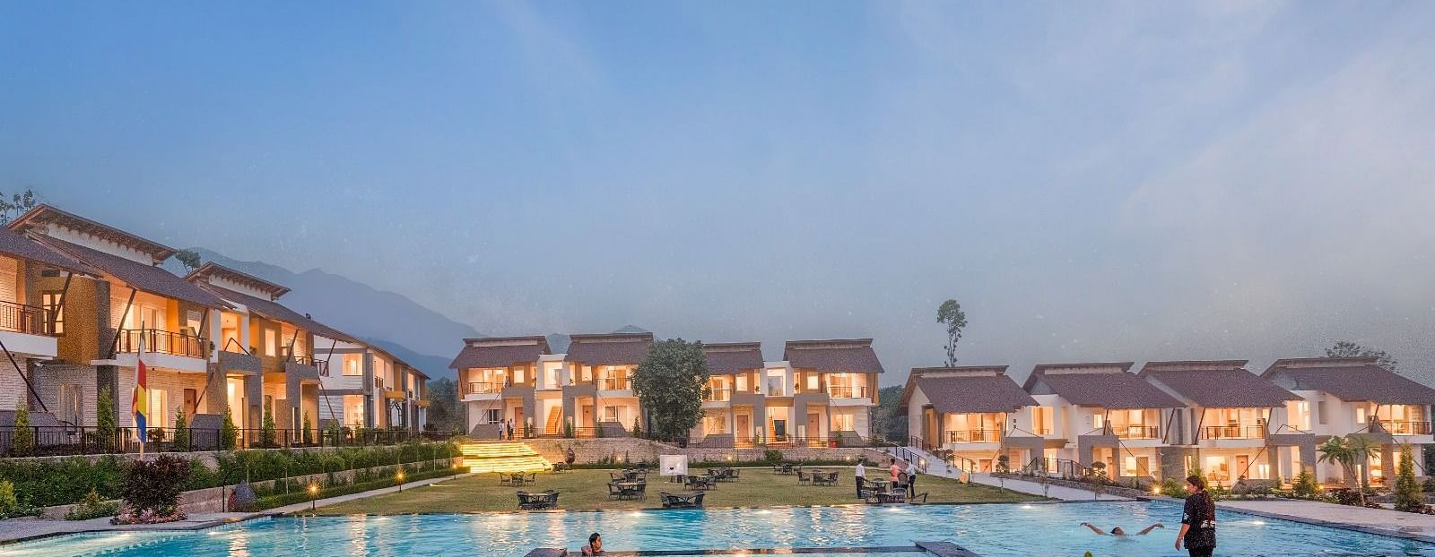 A large resort with multiple buildings surrounding a swimming pool, under a twilight sky.